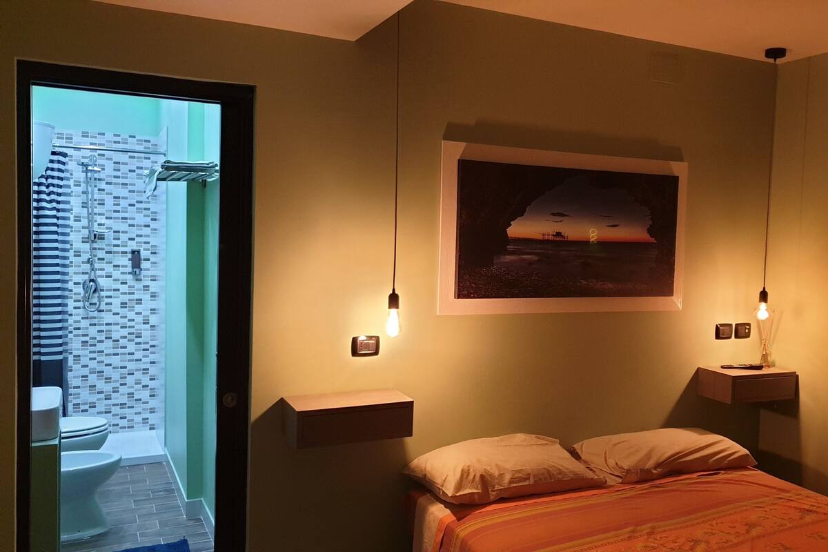 A bedroom with a bed, a nightstand, a wall-mounted lamp, a framed picture, and a view of a bathroom with a tiled wall and a shower curtain.