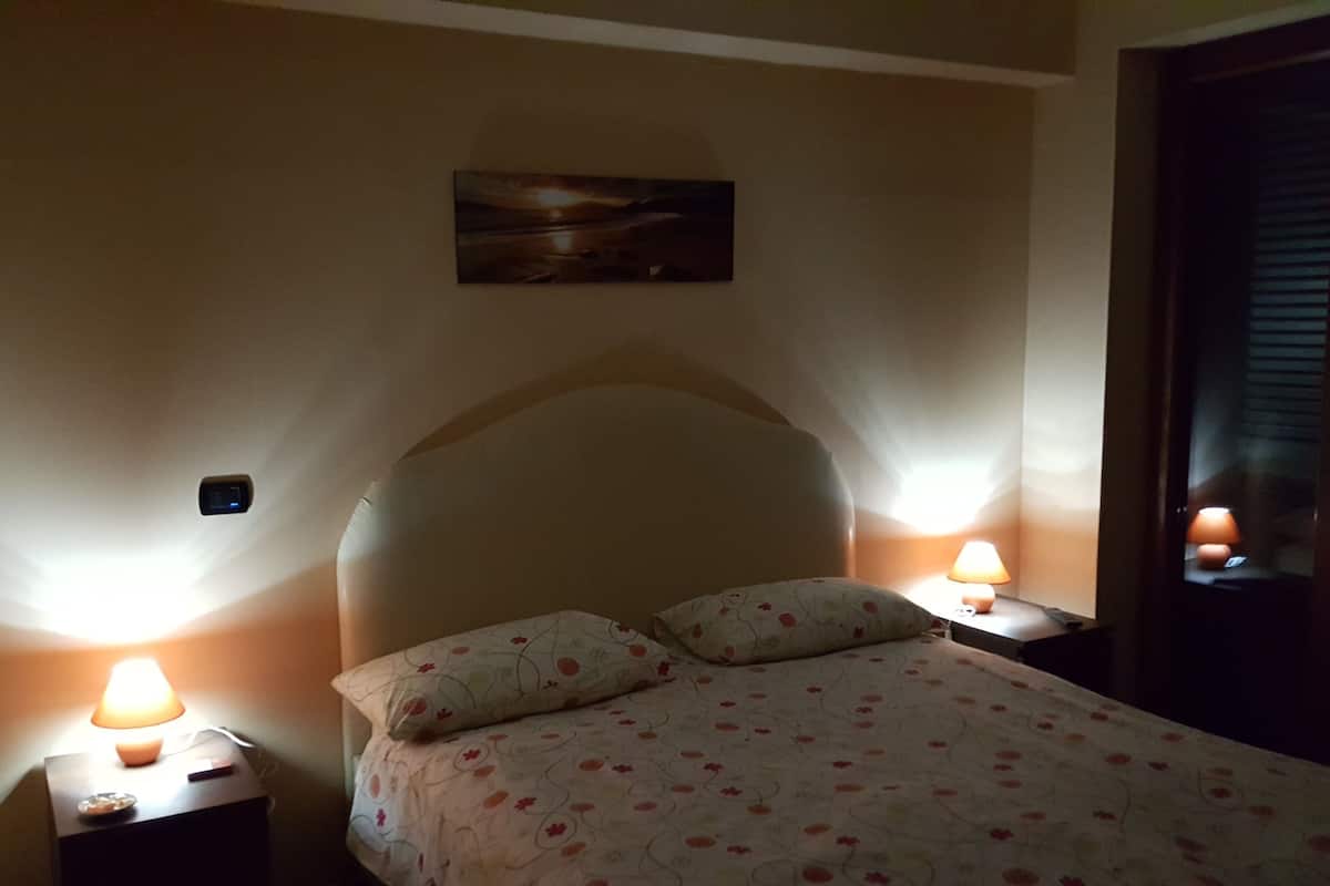 A bedroom with a bed, two bedside lamps, a nightstand, a wall clock, and a painting above the bed.
