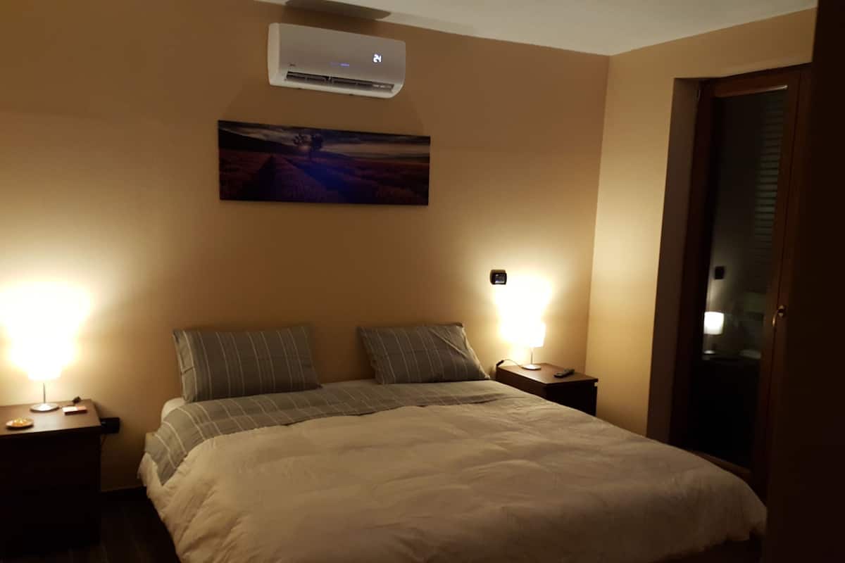 A hotel room with a bed, two bedside tables, a nightstand, a wall-mounted air conditioner, and a painting above the bed.