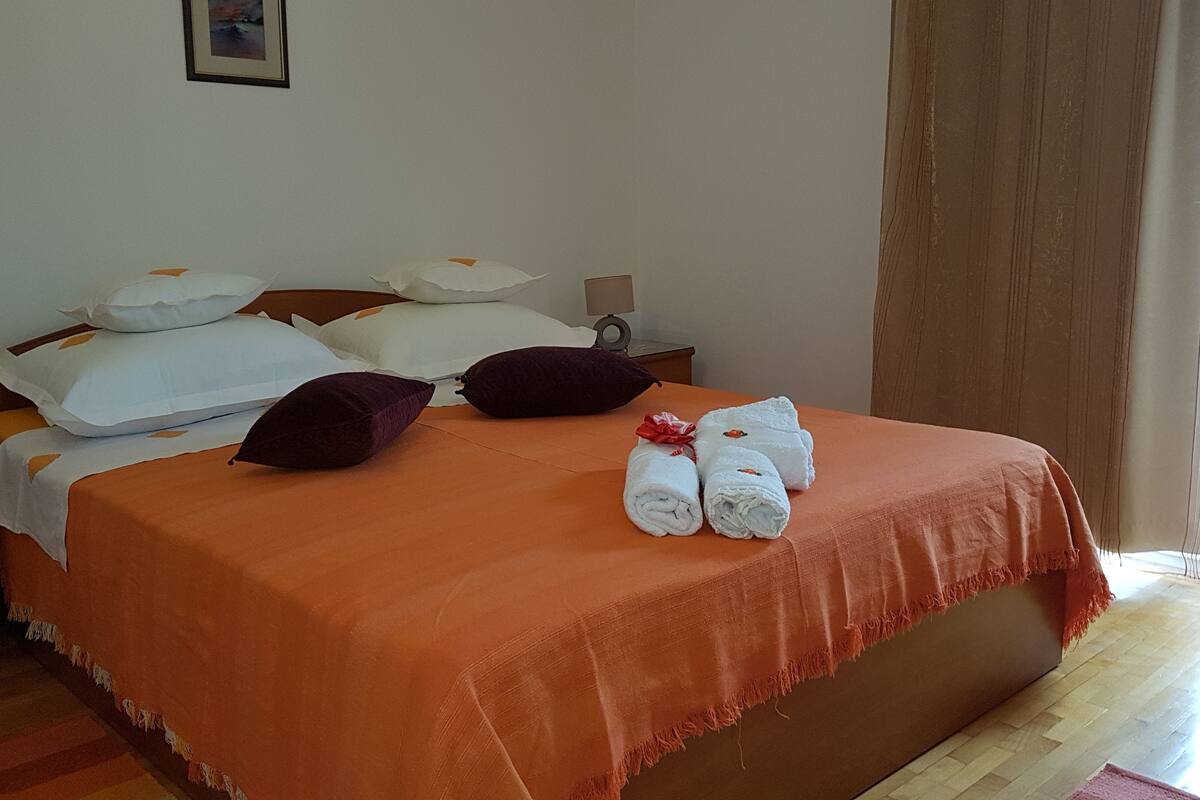Un lit bien fait, avec une couvre-lit orange, des oreillers blancs et une paire de serviettes enroulées, maintenues par un ruban rouge.