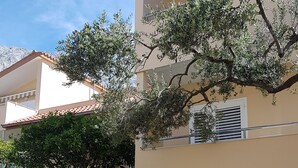 Ein Gebäude mit beige-farbener Fassade, einem Ziegeldach und einem Balkon. Vor dem Gebäude erstreckt sich ein Garten mit verschiedenen Blumen und einem großen Baum.