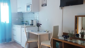 Eine kleine Küche mit weißen Schränken, einer Mikrowelle und einer Kaffeemaschine. Es gibt einen Esstisch mit zwei Stühlen und eine Vase mit Blumen. Ein Flachbildfernseher ist über dem Tisch an der Wand befestigt.