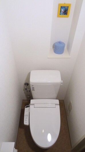 A small, compact bathroom with a white toilet, a wall-mounted shelf with a blue basket, and a plain white wall. - Family Lodge Hatagoya Matsuyama (Matsuyama)