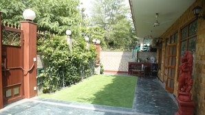 A courtyard with a green lawn, a red statue, a tiled floor, and a brick wall. - Dev Vatika Home Stay (Gurugram)