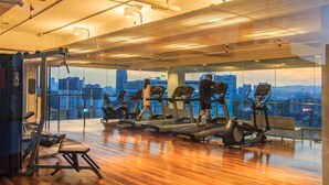 Een sportschool met loopbanden en trainingsapparatuur, grote ramen met stadszicht, en een houten vloer.