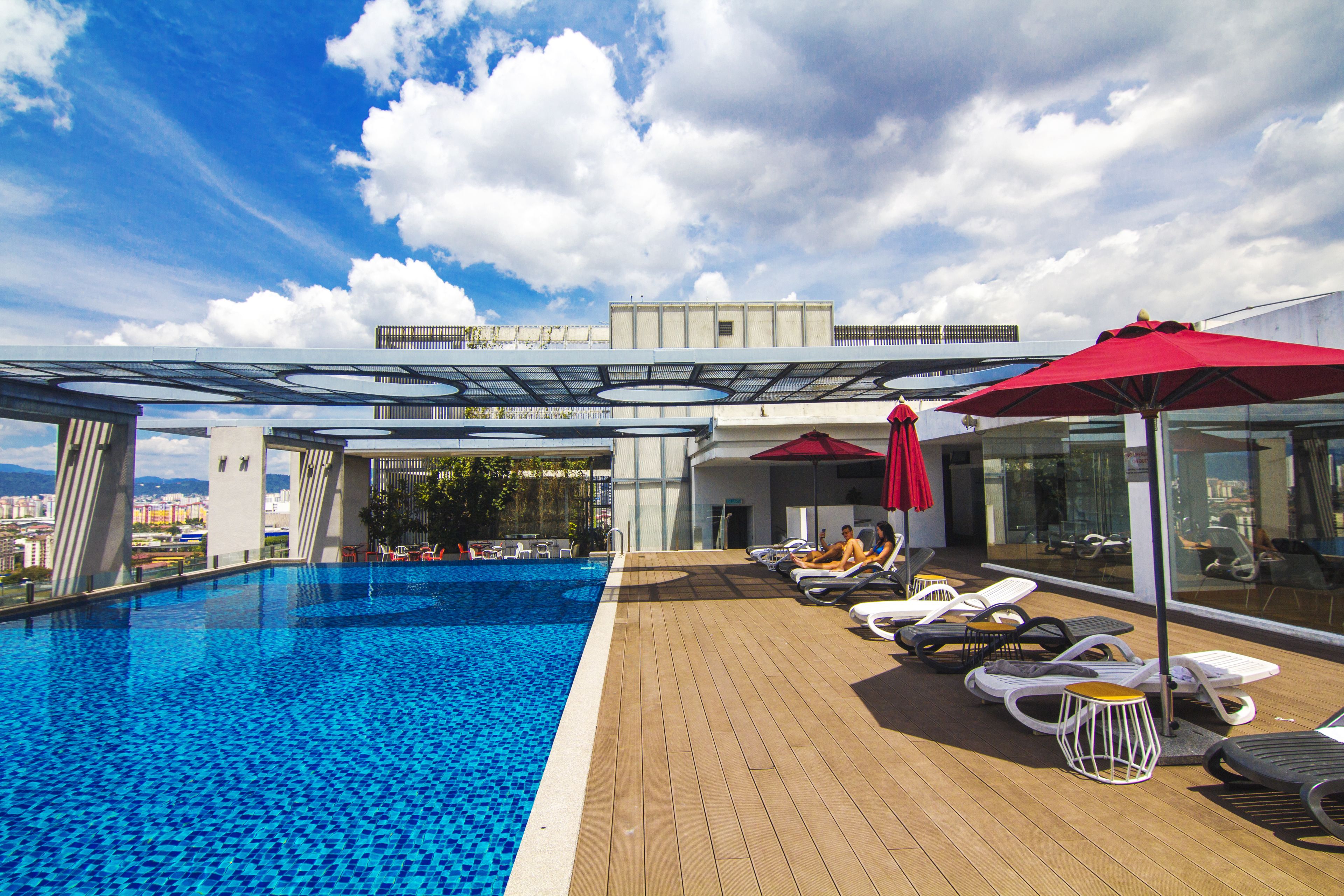 A rooftop swimming pool with lounge chairs and umbrellas, surrounded by a modern building with a glass facade.