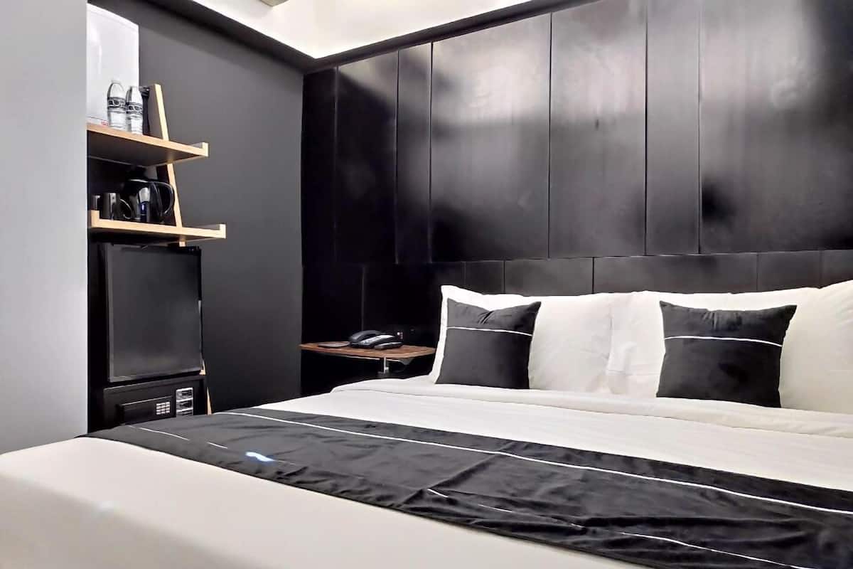 A modern hotel room with a large bed, a flat-screen TV, a bedside table with a phone, and a shelf with toiletries.