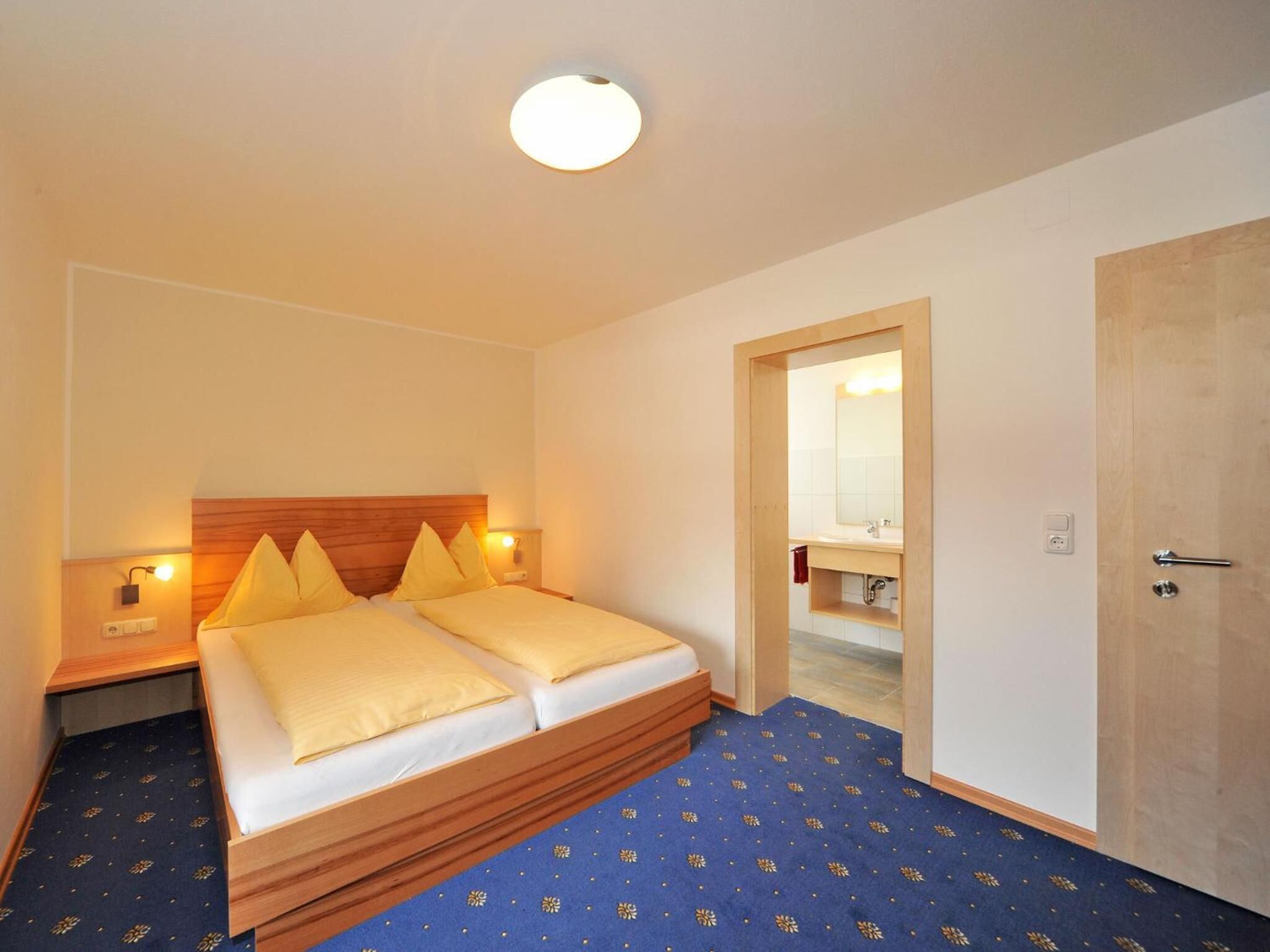 Een hotelkamer met een tweepersoonsbed, houten hoofdeinde, bedlampjes, een spiegel en een deur.