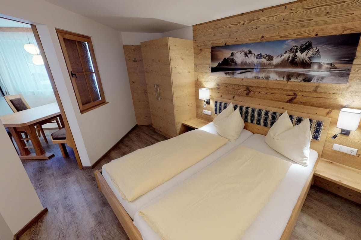 A bedroom with a wooden headboard, a painting of a mountain landscape, two beds, bedside lamps, and a wooden wardrobe.