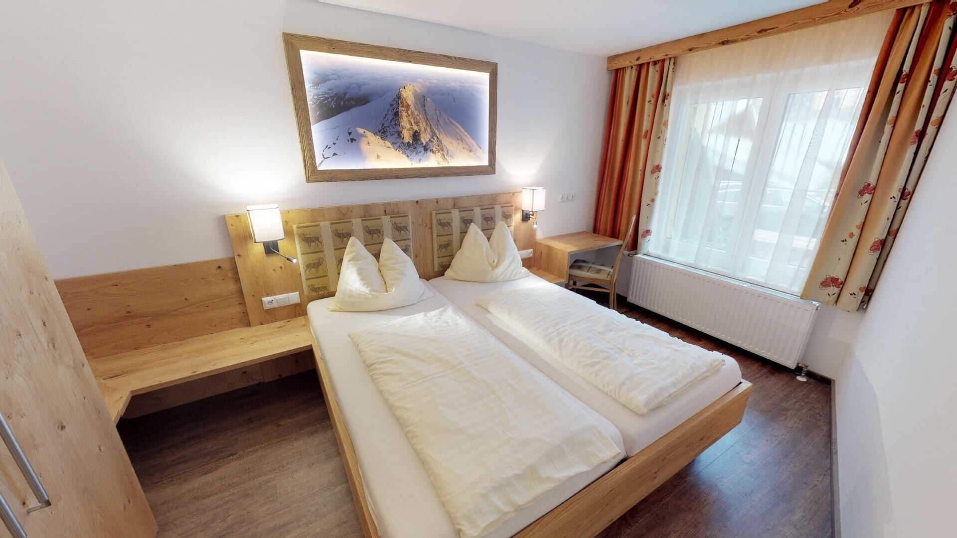 Hotelkamer met een houten bed, twee kussens, een nachtkastje, een bureau, een stoel, een raam met gordijnen en een ingelijst foto aan de muur.
