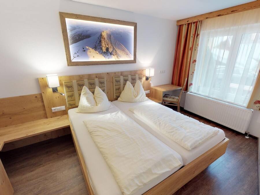 Hotelkamer met een houten bed, twee kussens, een nachtkastje, een bureau, een stoel, een raam met gordijnen en een ingelijst foto aan de muur.