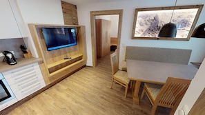 A modern kitchen with a built-in TV, wooden cabinetry, and a dining area with a view of mountains through a window. - Pension Bergkristall (Heiligenblut)