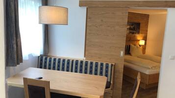 A compact living space with a wooden dining table, a bench with patterned cushions, a hanging lamp, and a sofa in the background.