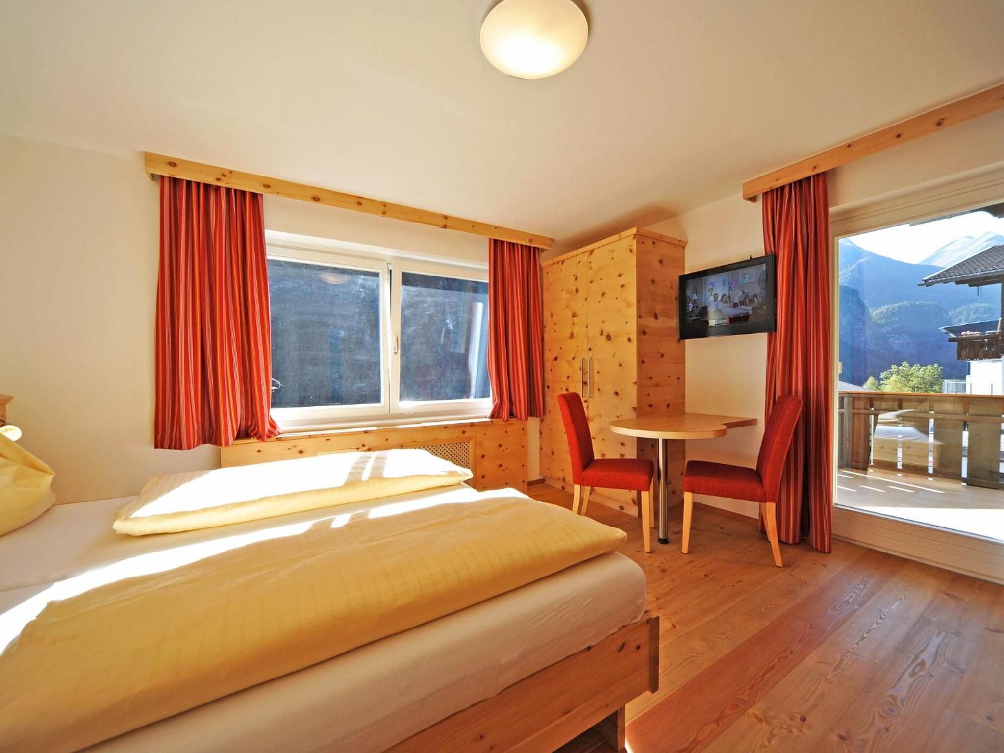 Una habitación con cama, una mesa, sillas, televisor y balcón con vista a la montaña.