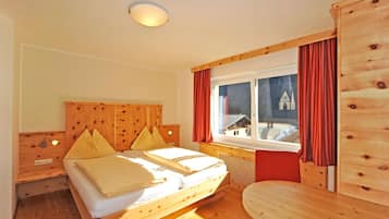 Een houten slaapkamer met een bed, nachtkastje, raam met gordijnen en uitzicht op een gebouw.