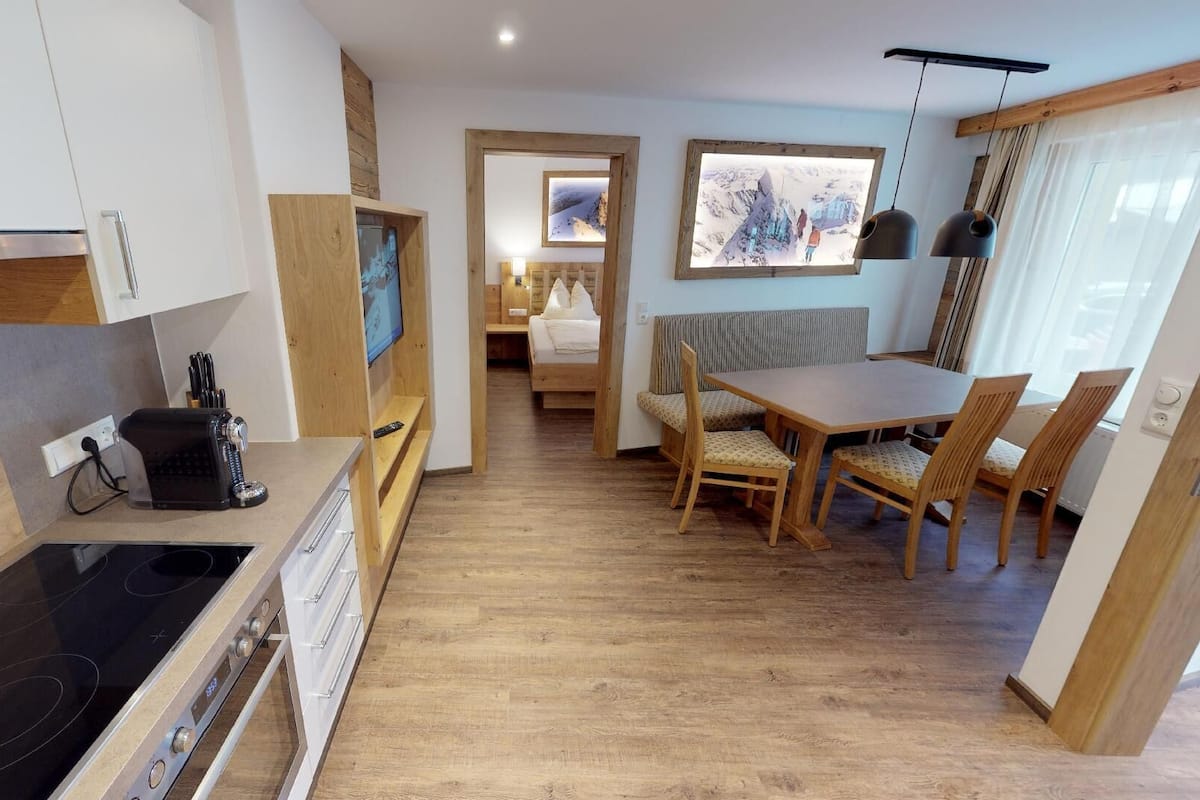 A modern kitchen with a stove, a dining area with wooden chairs, and a bed in the background.
