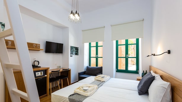 A hotel room with a bed, a desk with a computer, a TV, and a window with green shutters. - Ink Hotel Phos (Rethymno)