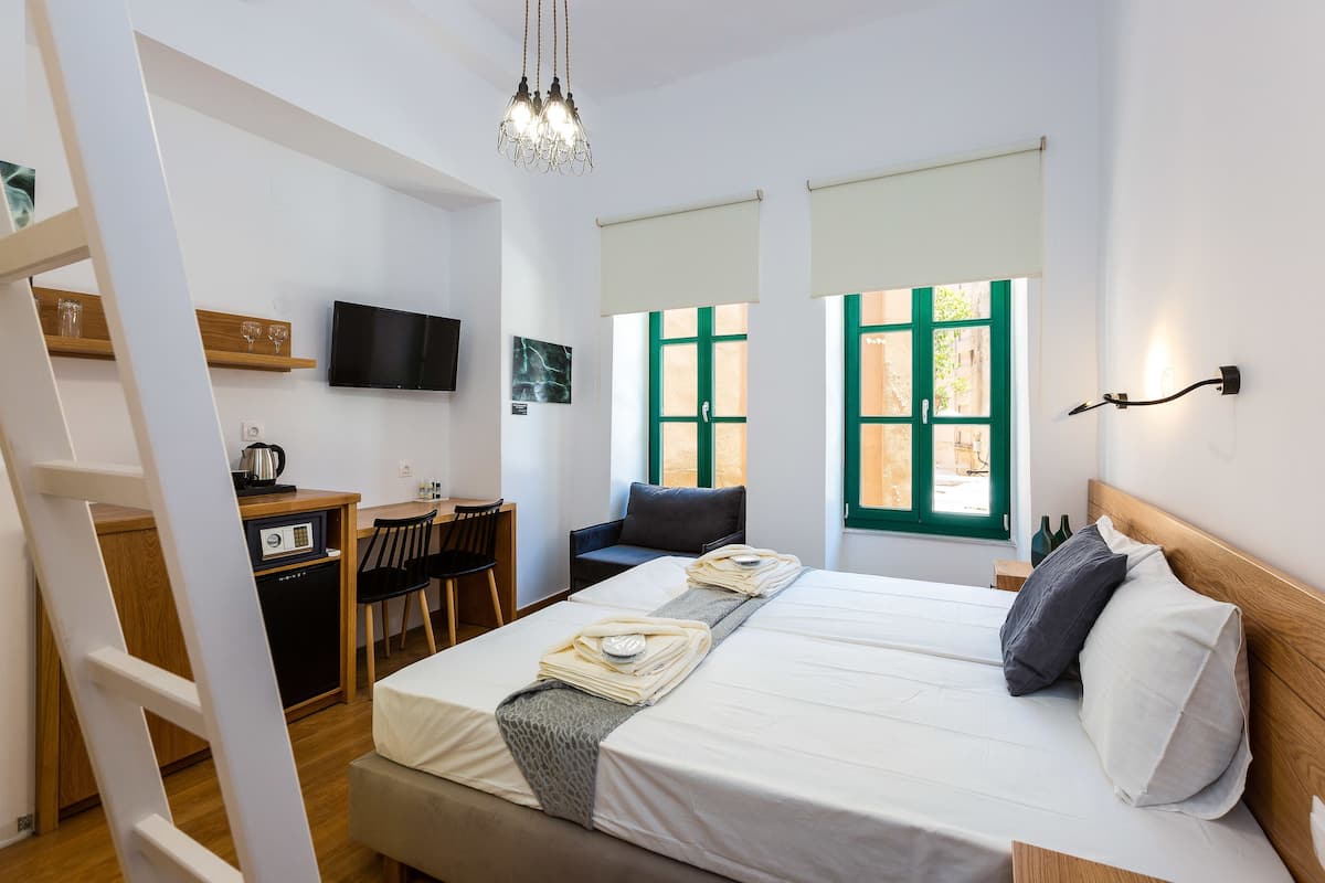 A hotel room with a bed, a desk with a computer, a TV, and a window with green shutters.