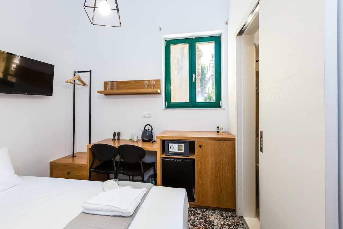 A hotel room with a bed, a desk with a chair, a small refrigerator, a TV mounted on the wall, and a window with a green frame.