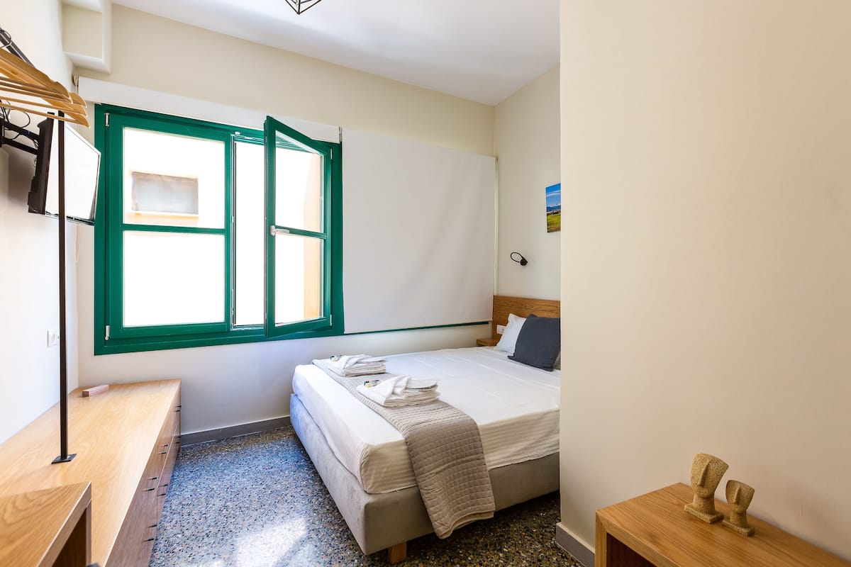 A bedroom with a bed, a wooden desk, a window with green frames, and a small wooden table with two decorative items.