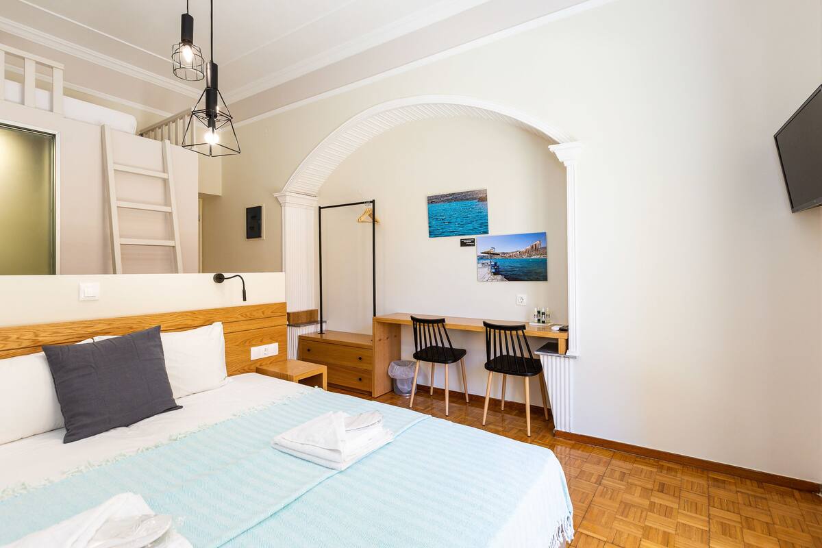 A bedroom with a bed, a desk, two chairs, a ladder, and a TV.
