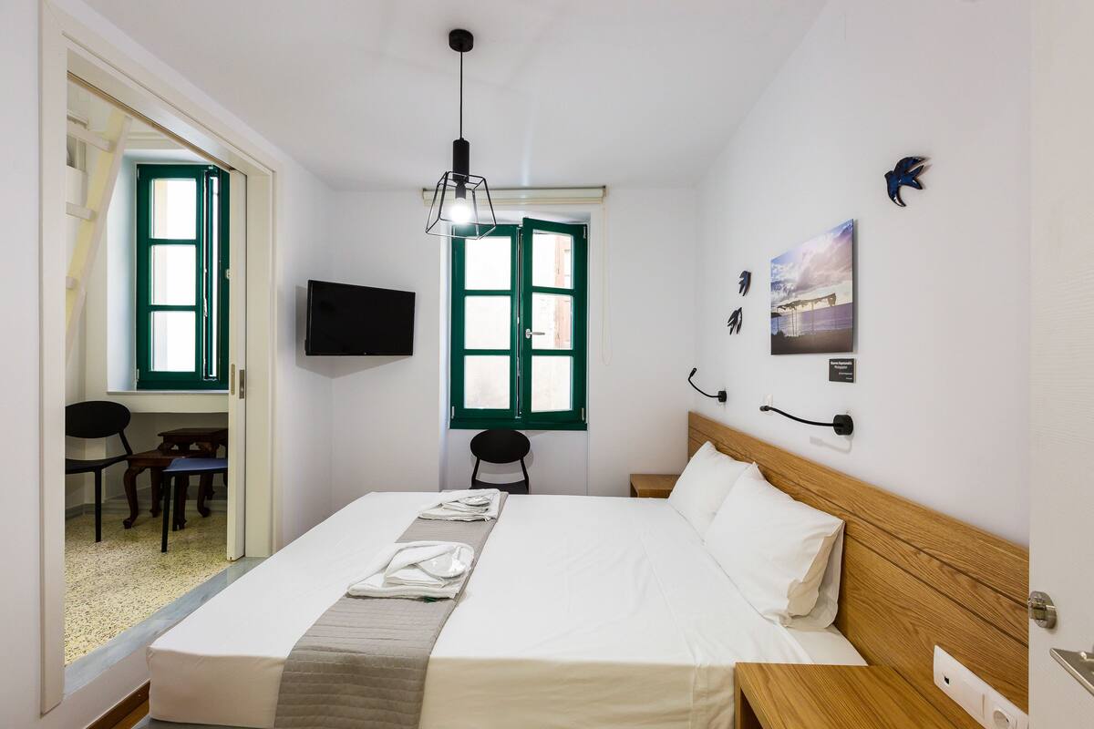 A hotel room with a bed, a desk, a chair, a television, and a window with green shutters.