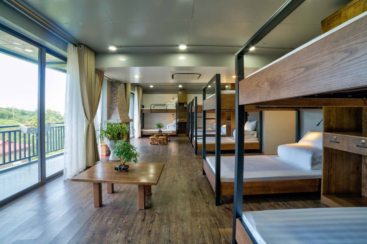 Ett modernt hotellrum med våningssängar, ett trägolv, ett stort fönster med gardiner, ett litet bord med en växt och en balkong med ett räcke.