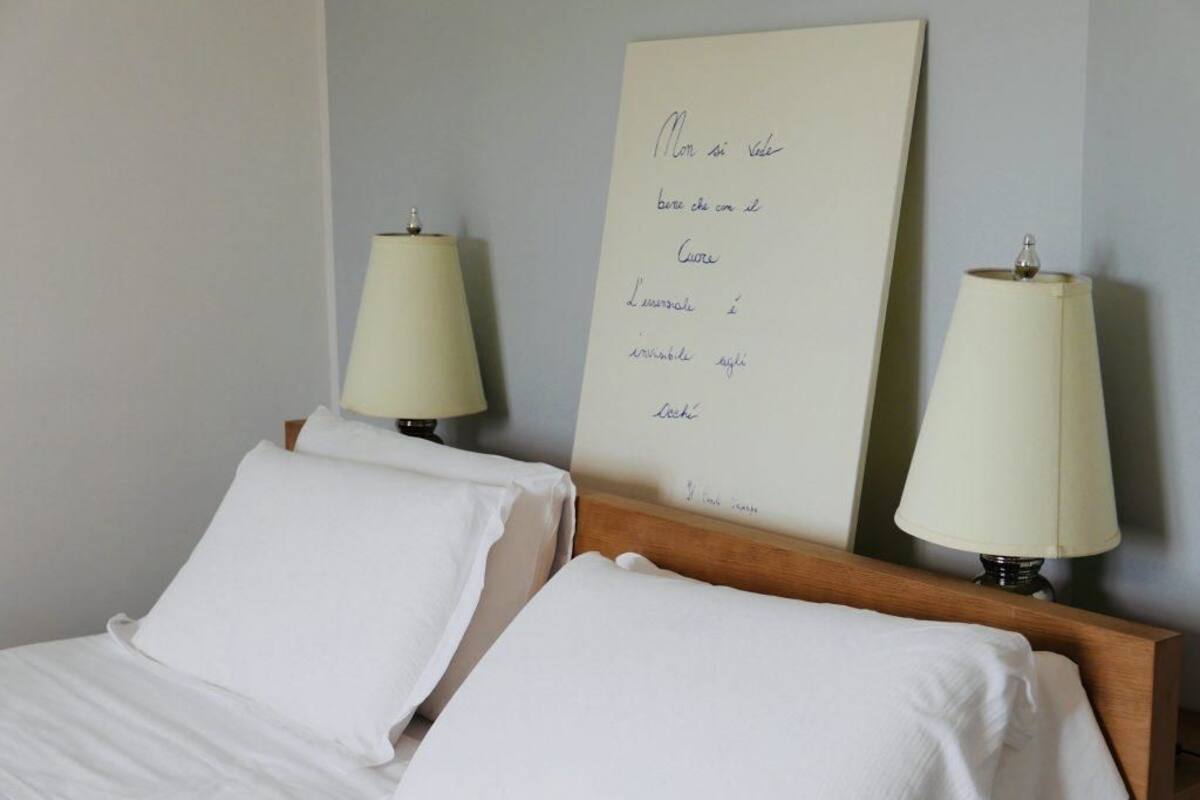 Una cama bien hecha con sábanas blancas, dos lámparas de noche y un letrero en la pared con texto manuscrito.