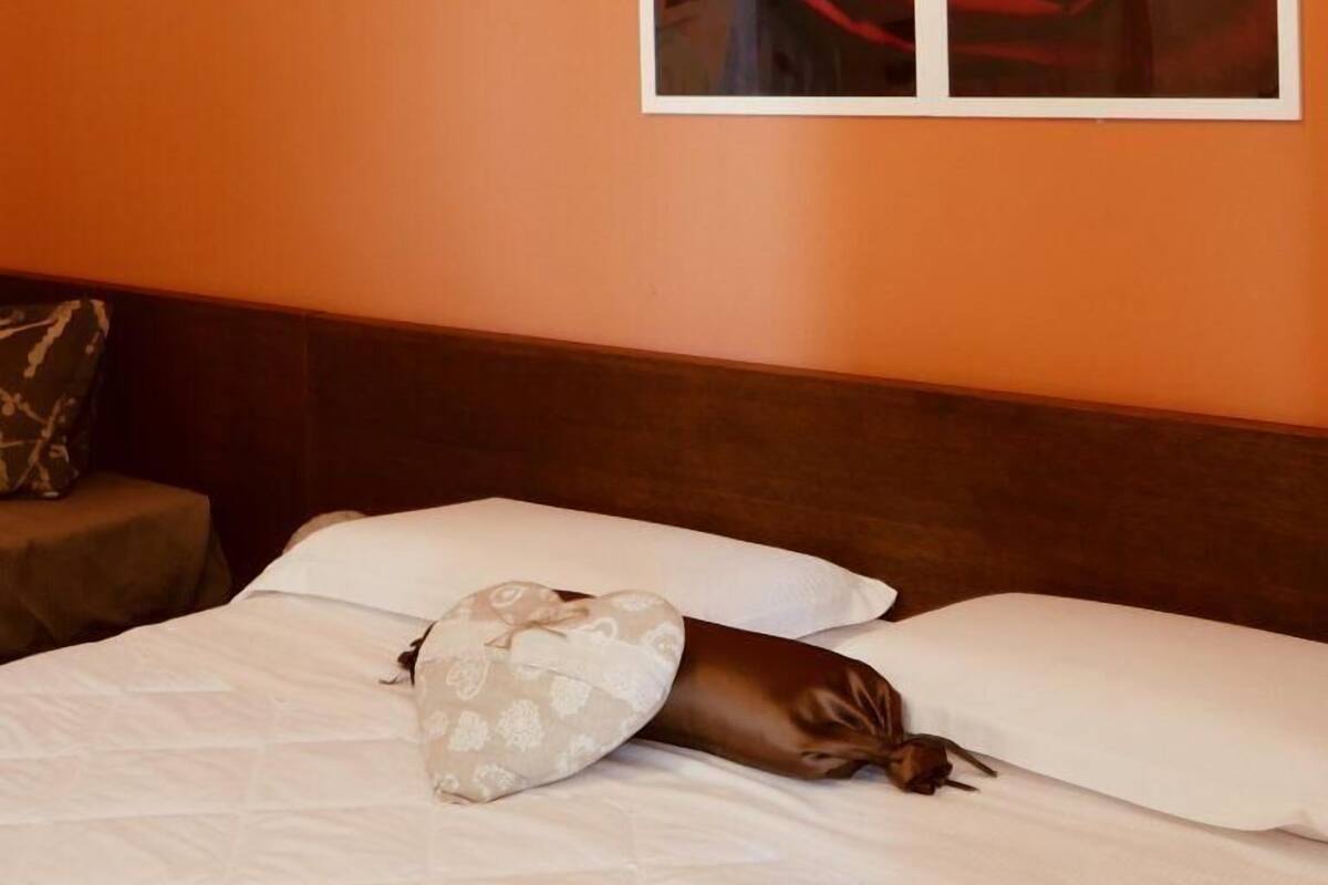 Habitación de hotel con pared naranja, ventana grande con pintura de una rosa roja, y cama con ropa blanca.