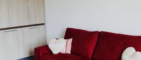 A red sofa with a white cushion and a heart-shaped pillow, a wooden wardrobe, and two framed landscape pictures on the wall.