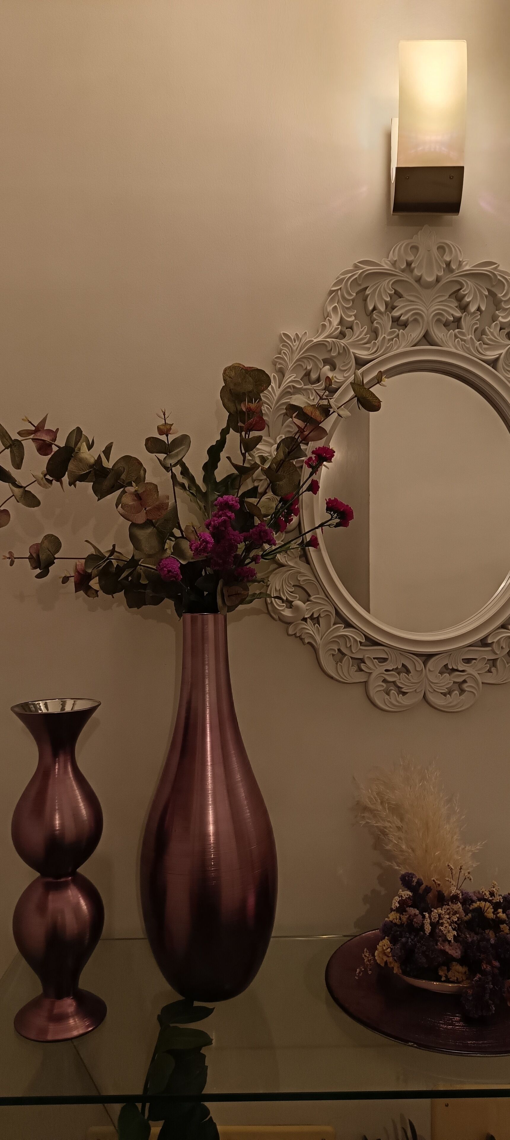 Eine dekorative Vase mit Blumen auf einem Tisch, ein Wandspiegel und eine Wandleuchte.