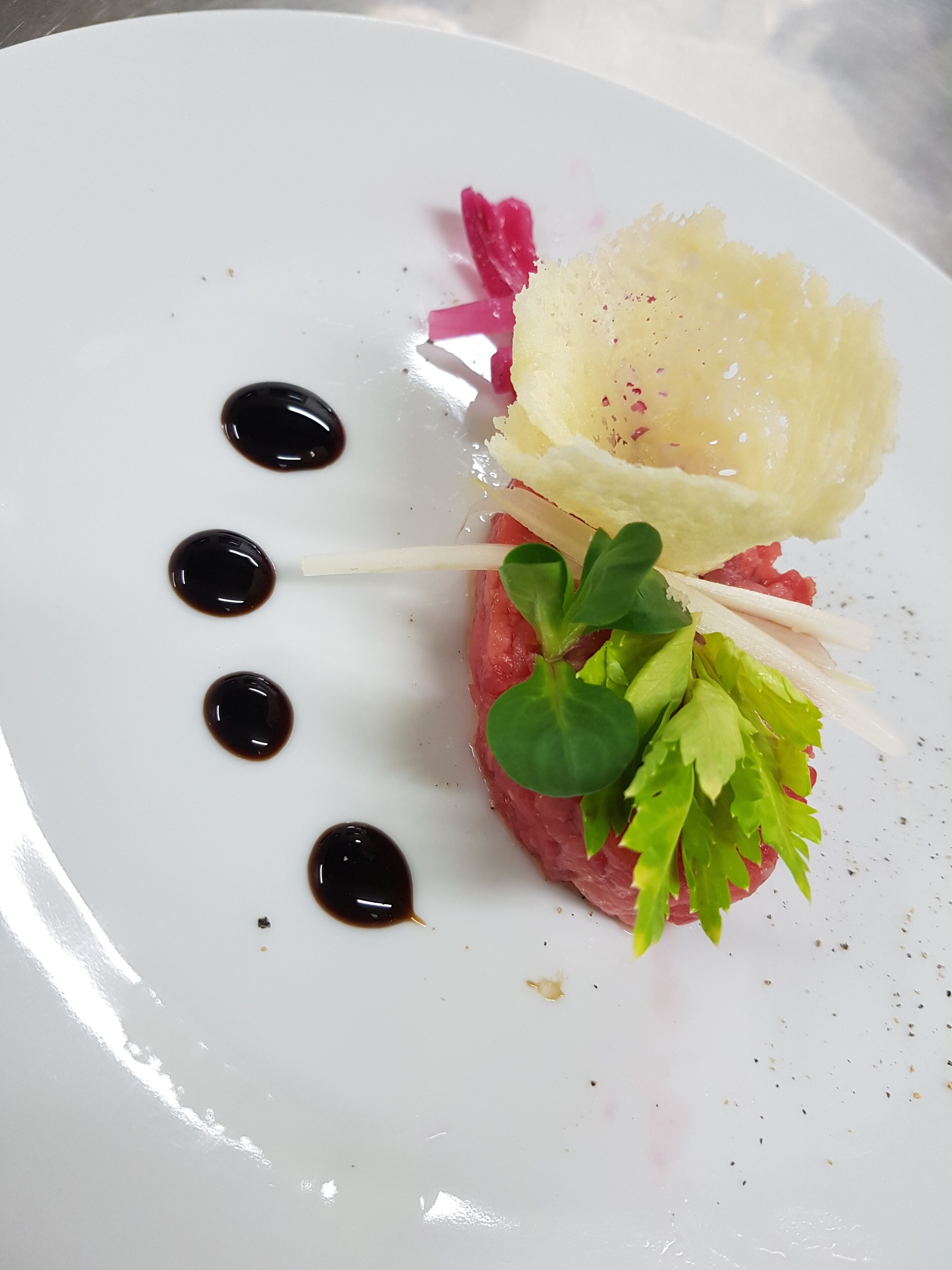 Un plato blanco con un guiso de carne cruda adornado con vegetales, una loncha de queso y una guarnición rosada.