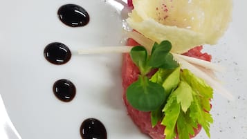 Un plato blanco con un guiso de carne cruda adornado con vegetales, una loncha de queso y una guarnición rosada.