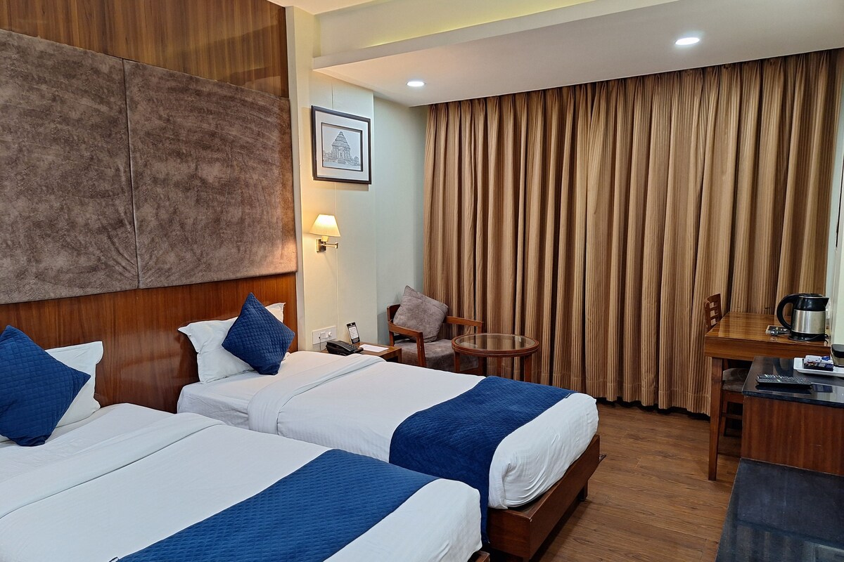 A hotel room with two beds, a desk, a chair, a lamp, and a large window with curtains.