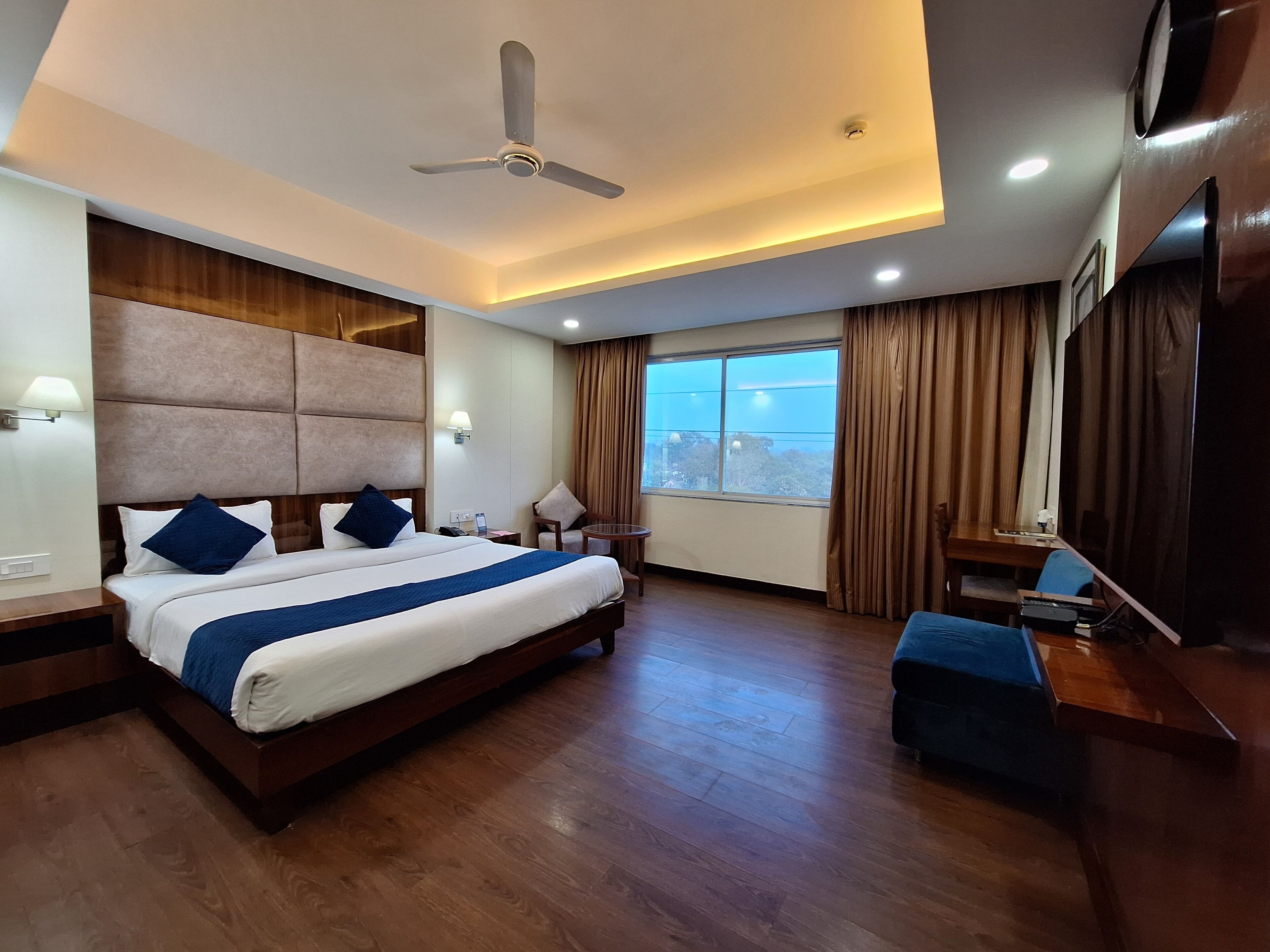 Une chambre d’hôtel avec un grand lit, un bureau, une chaise, un ventilateur de plafond et une grande fenêtre avec des rideaux.