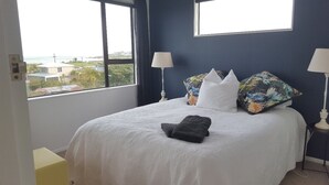 Una cama bien hecha con edredón blanco, dos almohadas y una toalla doblada. Hay dos lámparas de noche y una ventana con vistas a edificios y zonas verdes.