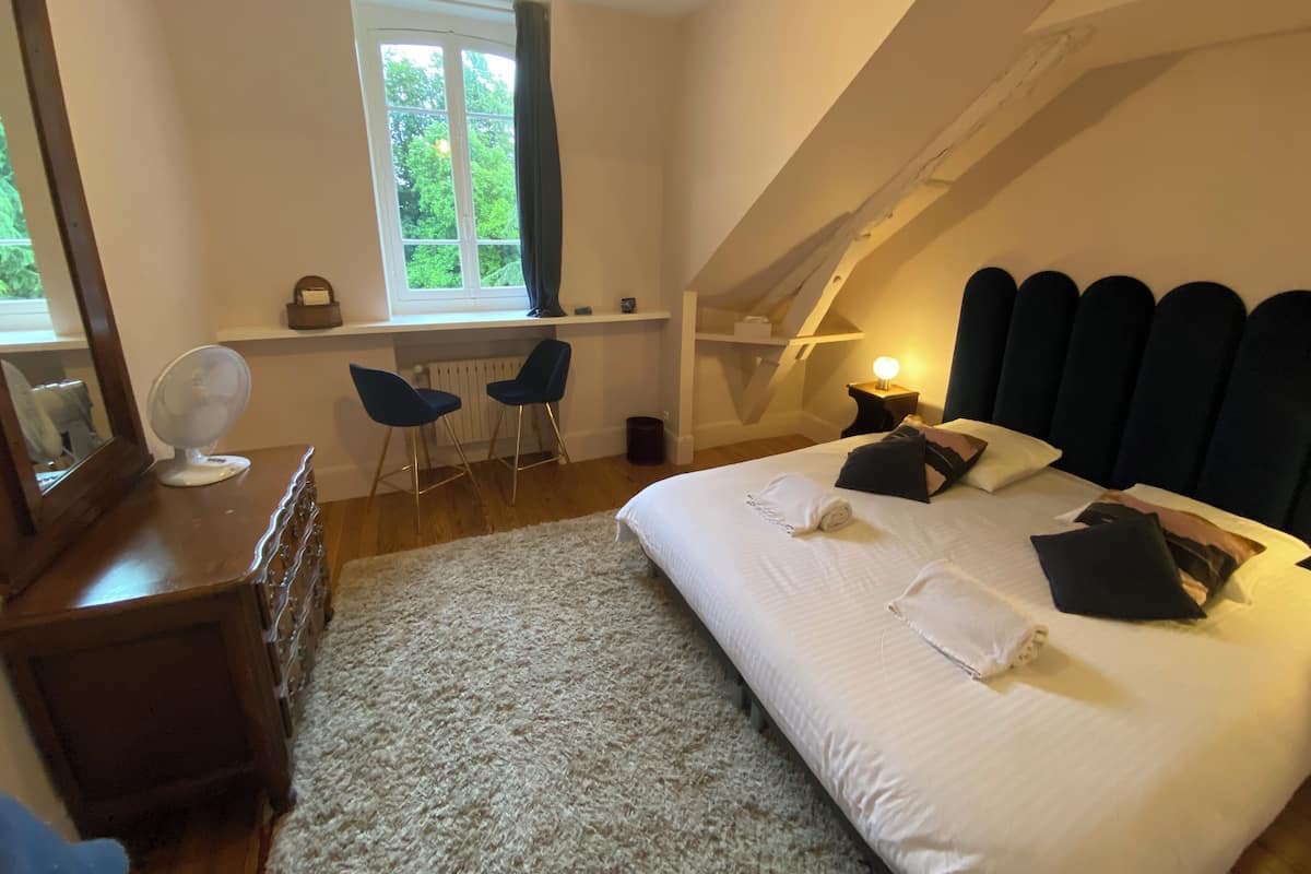 Een slaapkamer met een groot bed, een bureau met twee stoelen, een raam met gordijnen en een schuine plafond.