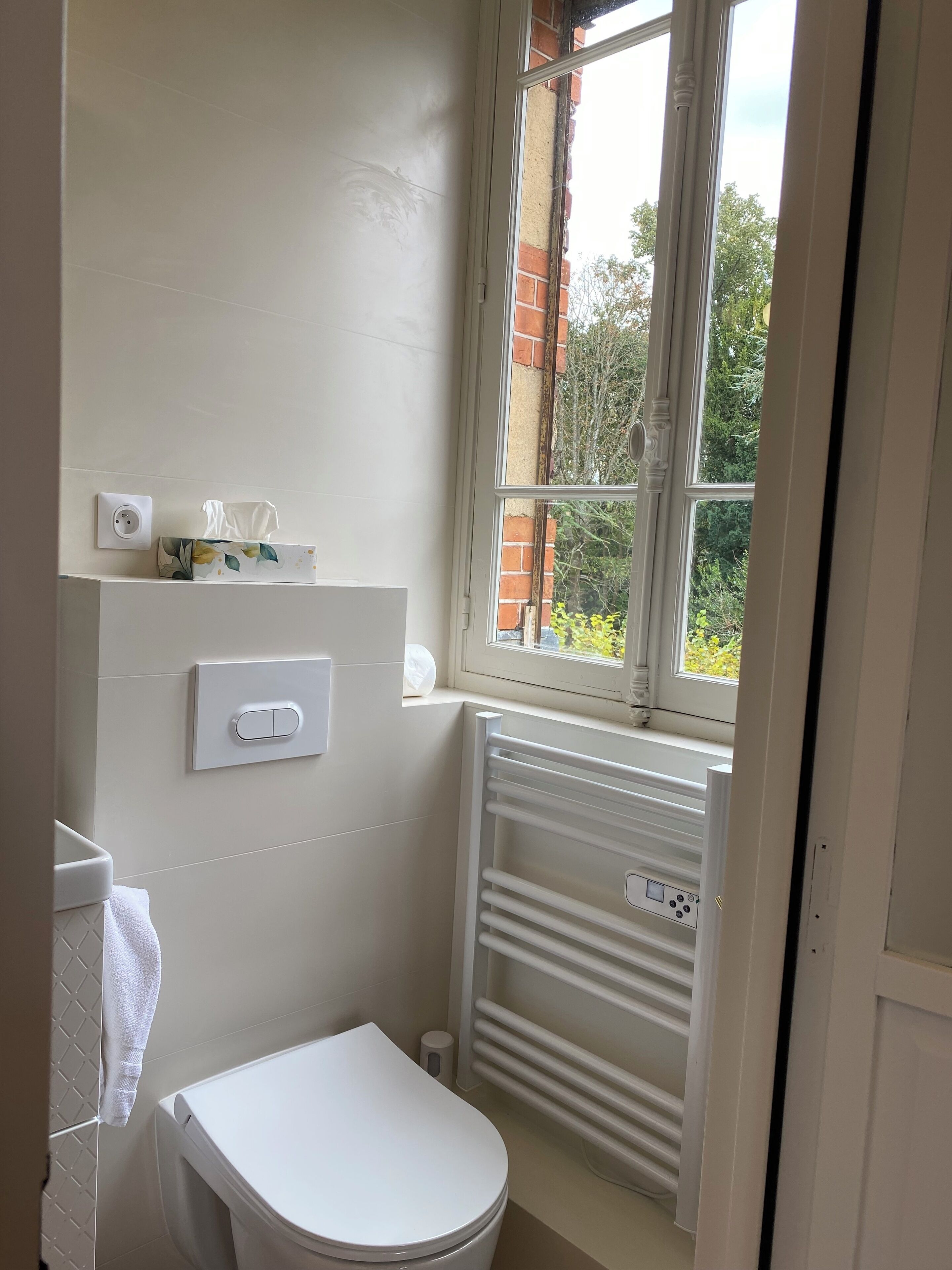 Une petite salle de bain propre, équipée de toilettes, d’un porte-serviettes, d’un radiateur et d’une fenêtre donnant sur un espace verdoyant.