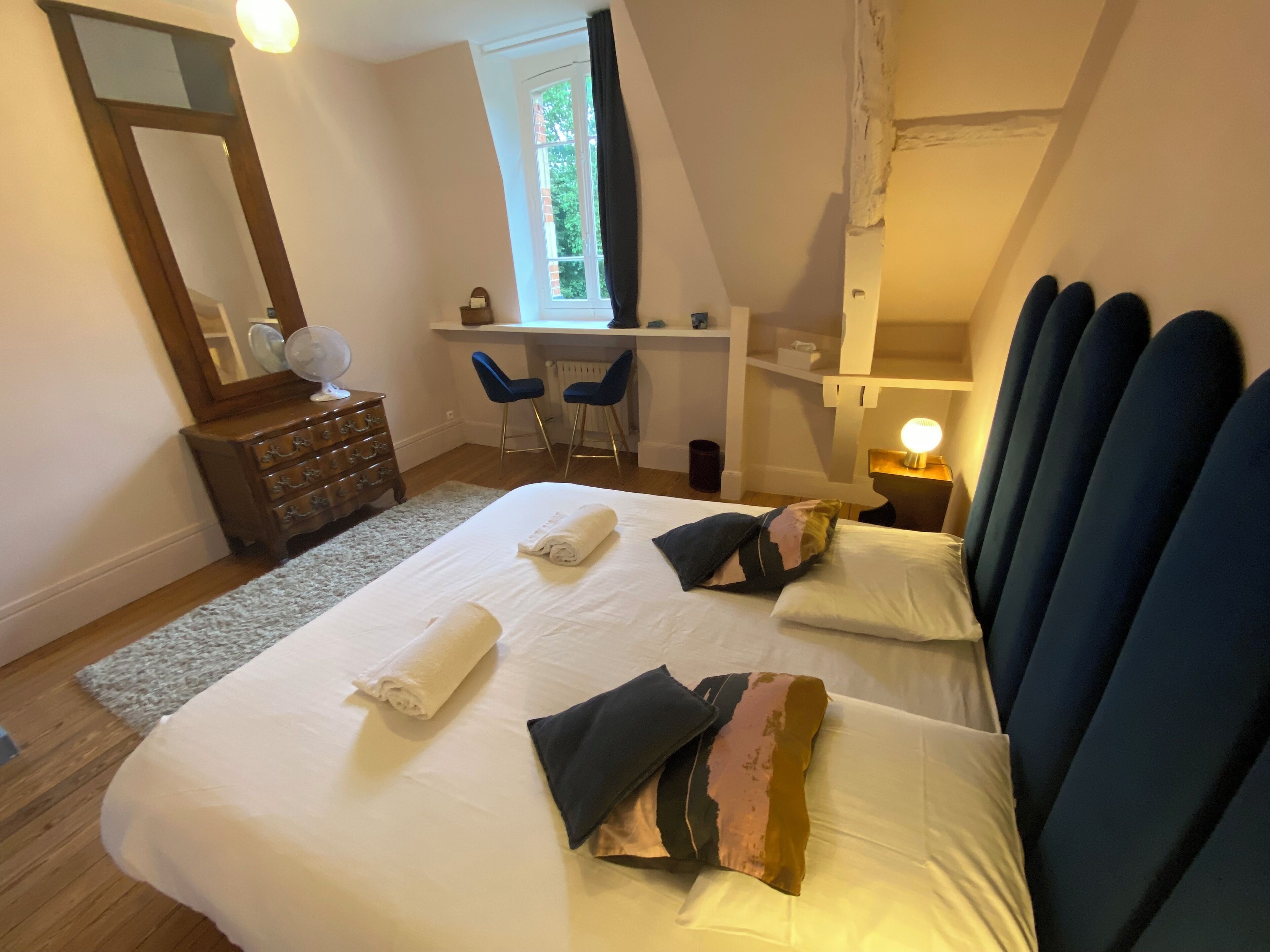 A bedroom with a large bed, a dresser, a desk with two chairs, a mirror, and a window with curtains.
