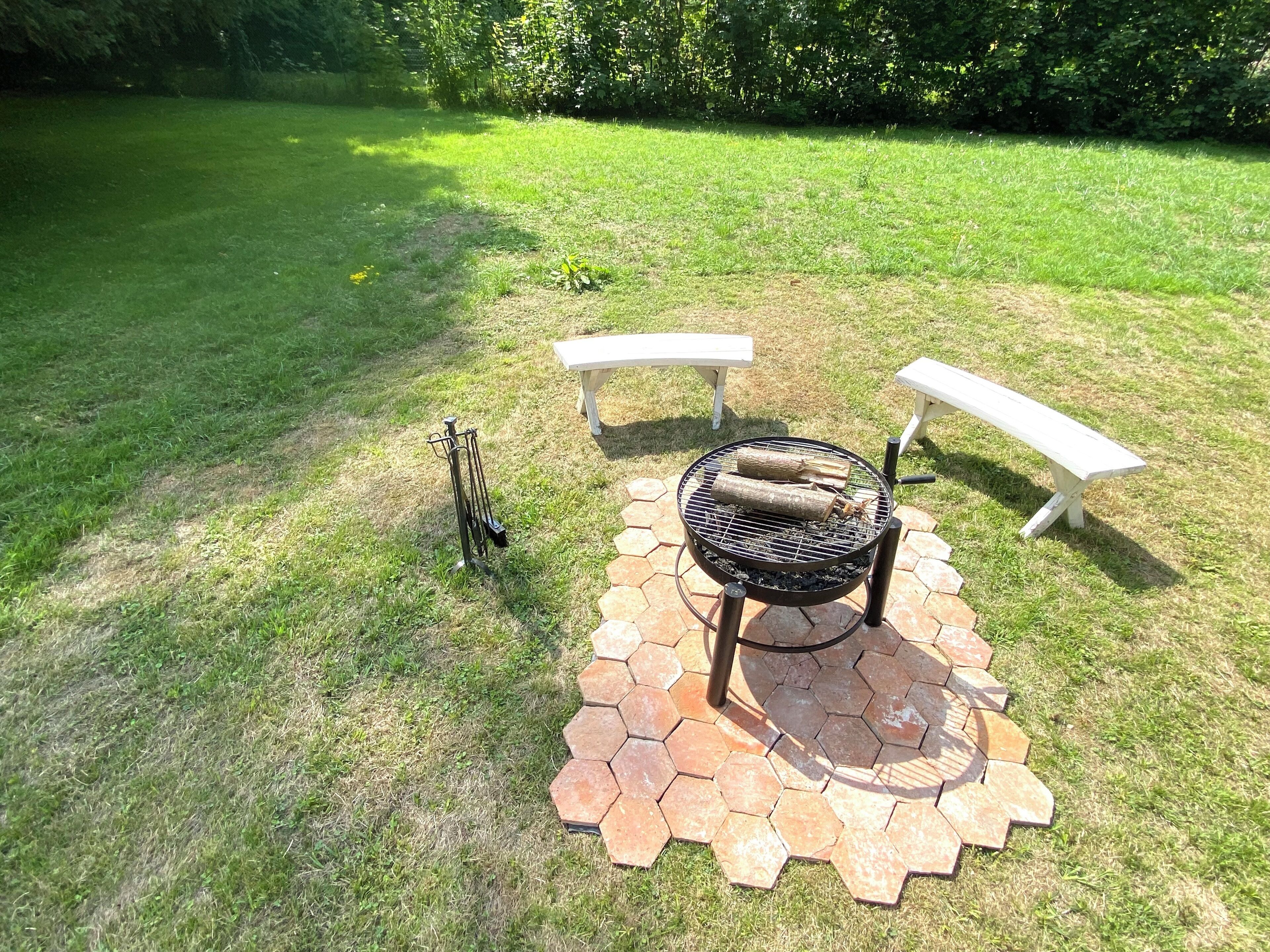 A barbecue grill with hexagonal tiles, two white benches, and a metal grill stand in a grassy outdoor area.