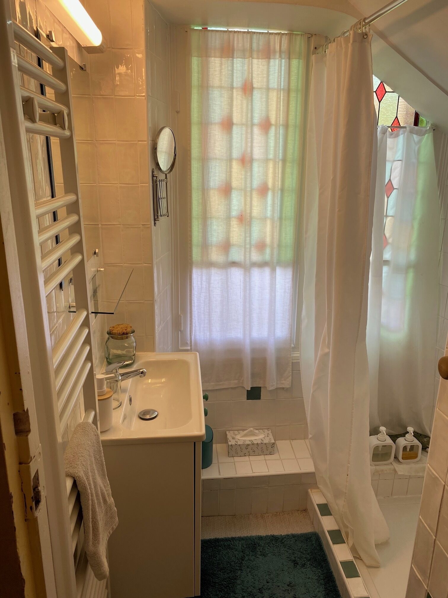 Un baño con un lavamanos blanco, un espejo, un perchero para toallas, una cortina de ducha y una ventana con vidrieras.
