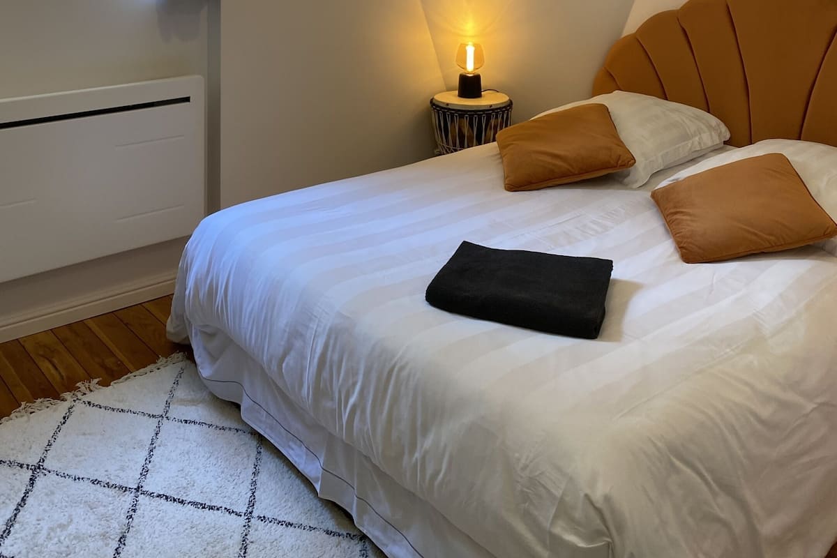 Een slaapkamer met een bed, kussens, een bedlampje, een raam met zonwering en een radiator.