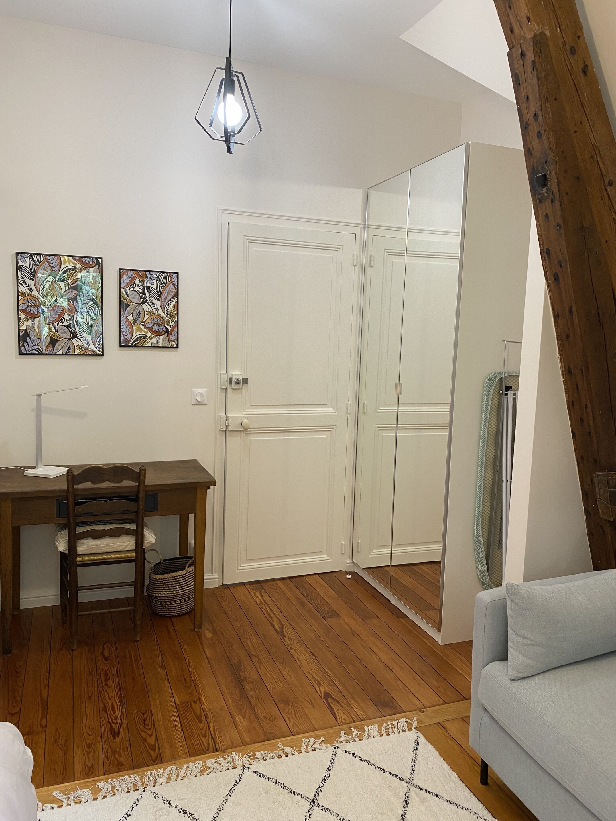 Una habitación con un escritorio de madera, dos obras de arte enmarcadas, una silla, un sofá, un espejo y una lámpara colgante.