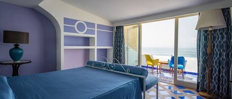 Habitación de hotel con cama, una mesa con lámpara, una silla y vista a la playa.
