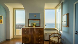 A room with a wooden dresser, a chair, a large window with a view of the sea, and a framed picture on the wall.