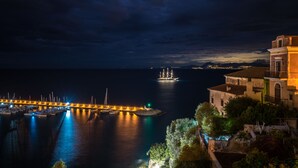 A coastal marina at night with boats docked, a distant ship, and buildings with balconies overlooking the water.