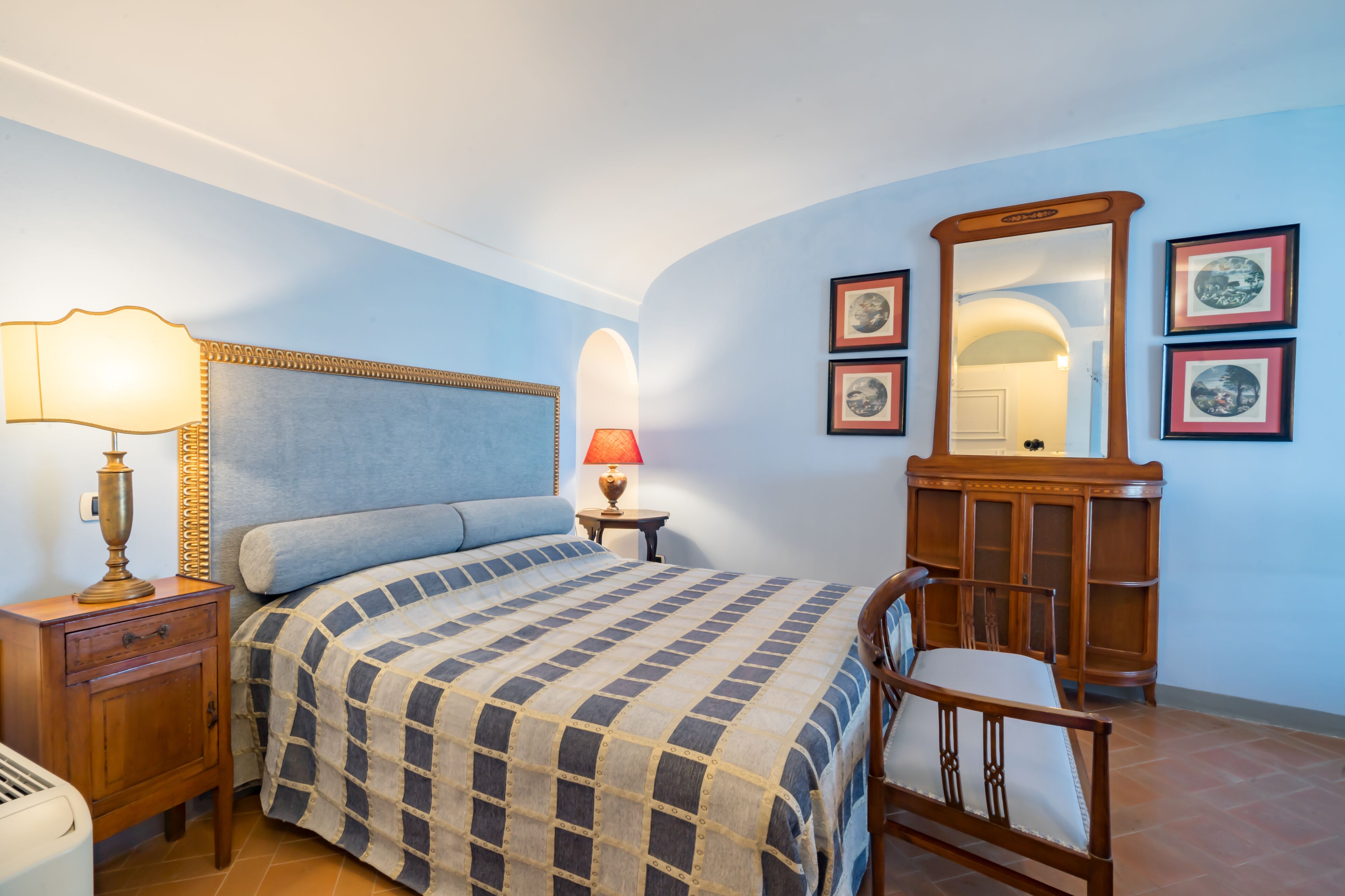 Un dormitorio con cama, mesita de noche, lámpara, cómoda, silla e imágenes enmarcadas en la pared.