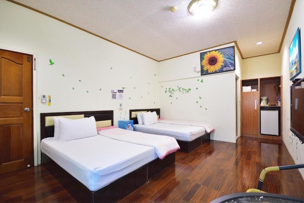 A hotel room with three beds, a wooden floor, a ceiling light, and a wall with a sunflower decoration.