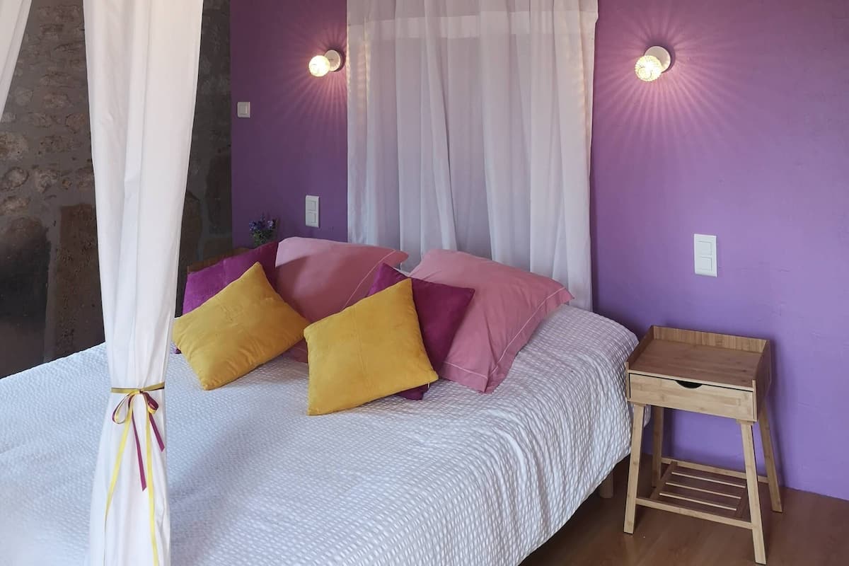 Een slaapkamer met een bed, witte gordijnen, paarse muren en een houten plafond.