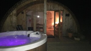 Una cabaña de madera con un jacuzzi en primer plano y un interior acogedor visible a través de la puerta abierta.